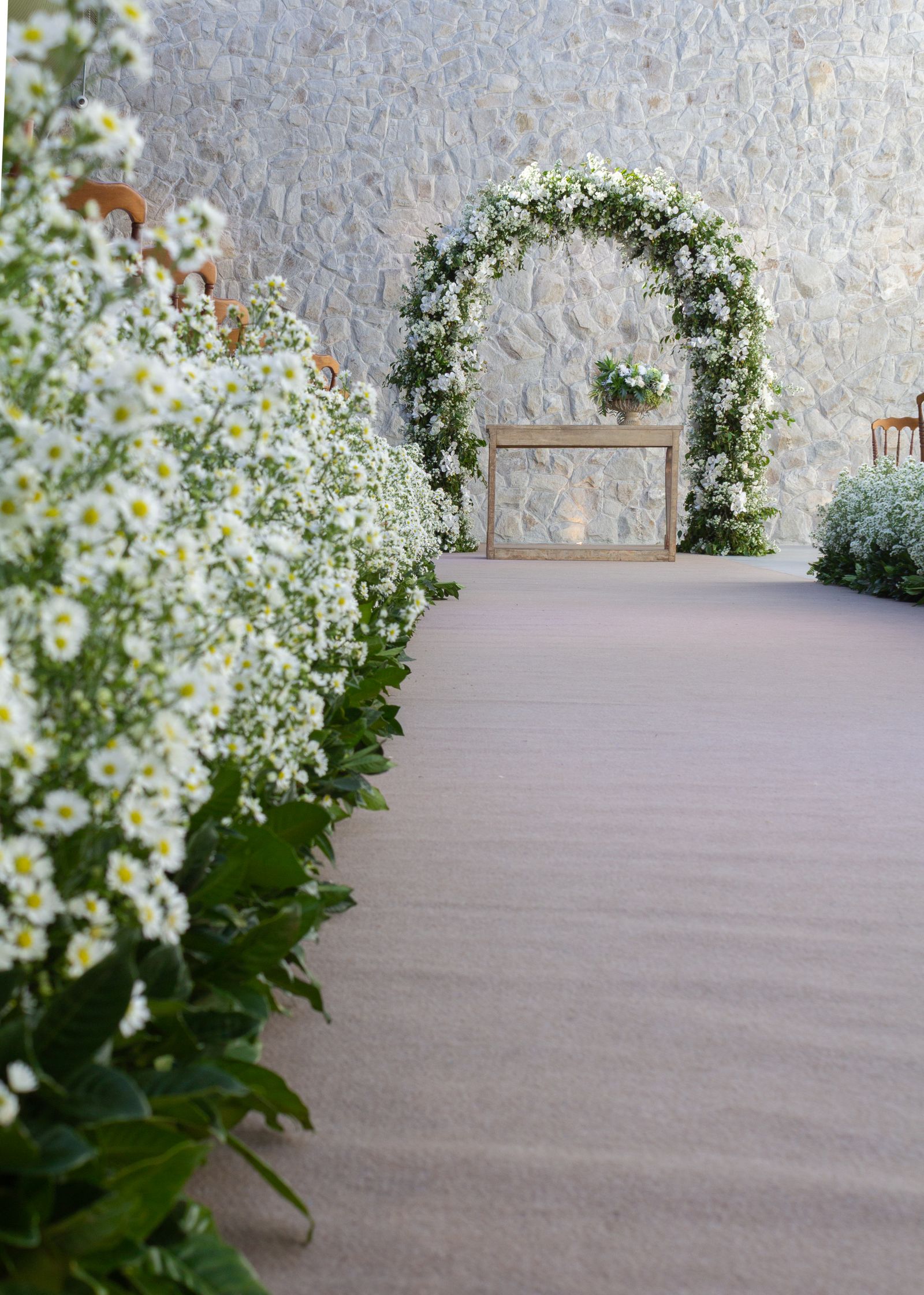 White floral wedding arch and flower-lined aisle against a stone wall